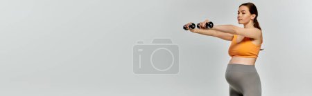 A pregnant woman in activewear confidently lifts dumbbells in front of a white background.