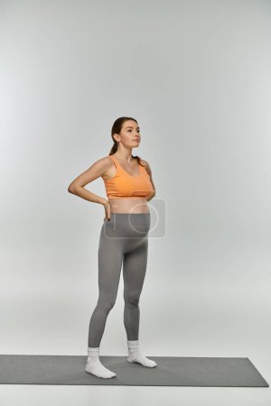 Eine sportliche Schwangere in Trainingskleidung steht selbstbewusst auf einer Matte, die Hände auf den Hüften und zeigt ihre Stärke und Vitalität.