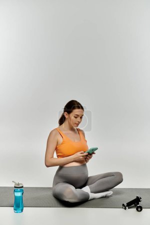 Sportliche Schwangere in Aktivkleidung sitzt auf Yogamatte und checkt Handy.