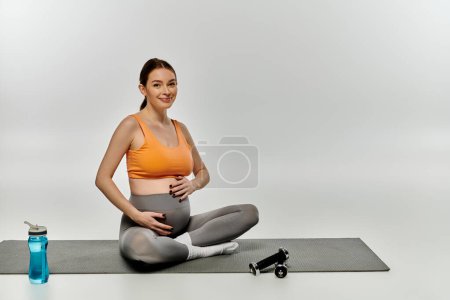 Eine schwangere Frau in aktiver Kleidung sitzt auf einer Yogamatte neben einer Wasserflasche.