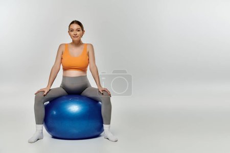 A young, pregnant woman in activewear balances gracefully on top of a vibrant blue exercise ball.