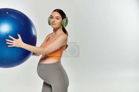 A pregnant, sporty woman in active wear balances a large blue ball in one hand while holding headphones in the other.