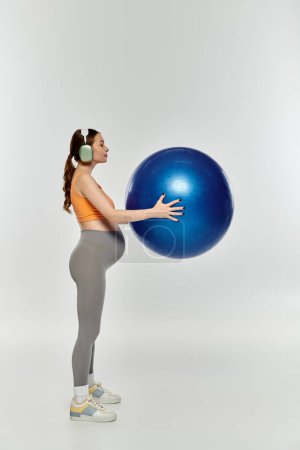 A young, sporty pregnant woman in activewear holds a large blue ball on a grey background.