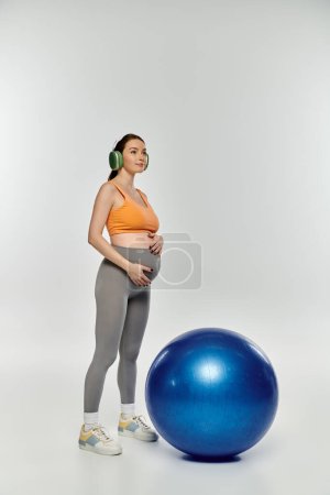 Eine junge, sportliche Schwangere in aktiver Kleidung steht anmutig neben einem blauen Trainingsball auf grauem Hintergrund..