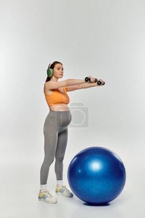 Sportliche Schwangere in aktiver Kleidung beim Training auf dem Gymnastikball mit Kopfhörern auf grauem Hintergrund.