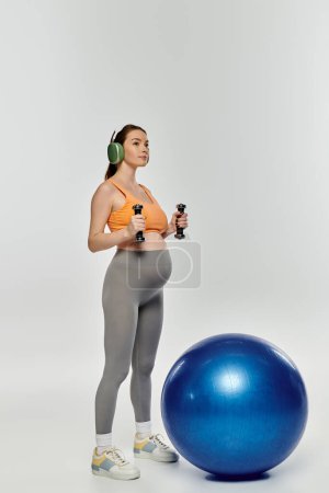 A sporty, pregnant woman stands next to a bright blue exercise ball against a grey background.