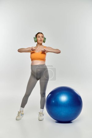 Foto de Una mujer embarazada en ropa deportiva se para junto a una bola de ejercicio azul brillante sobre un fondo gris. - Imagen libre de derechos