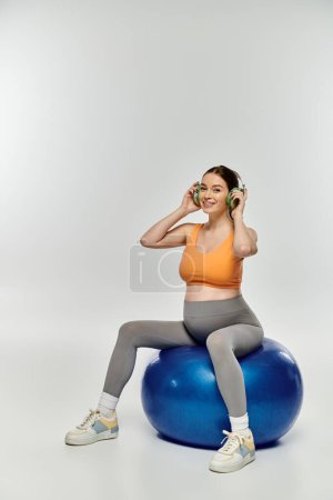 Foto de Young pregnant woman in active wear balances on blue ball, deeply engaged in music. - Imagen libre de derechos