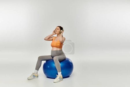 Eine junge, sportliche Schwangere in Aktivkleidung und Kopfhörer sitzt auf einem Turnball.