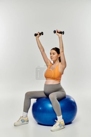 A young, pregnant woman in activewear sits on an exercise ball, holding two dumbbells, working out on a grey background.