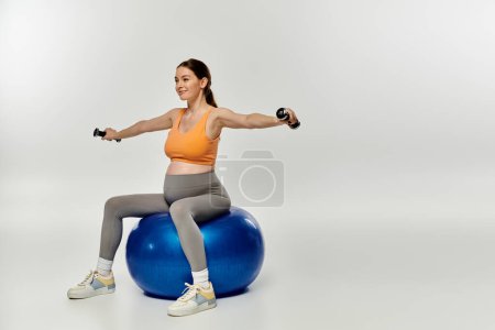 A pregnant woman in athletic attire sits on a stability ball while holding a dumbbell.