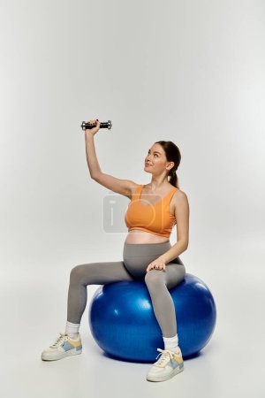 A pregnant woman in activewear demonstrates balance by sitting on a ball while holding a dumbbell.
