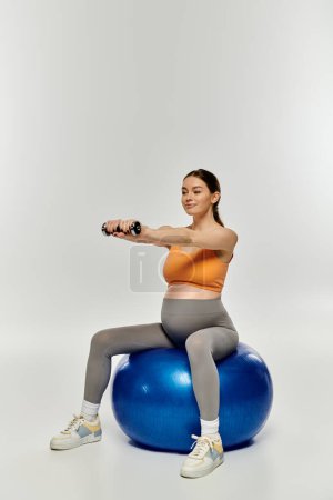A pregnant woman in activewear sits on an exercise ball, balancing with focus.