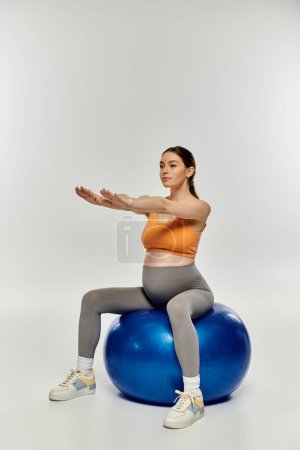 A young, pregnant woman in active wear sits balanced on a blue exercise ball, displaying strength and focus.
