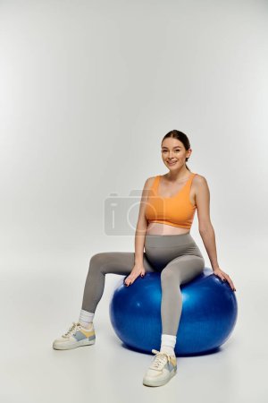 A young, sporty pregnant woman in active wear sits gracefully on a bright blue exercise ball against a neutral backdrop.