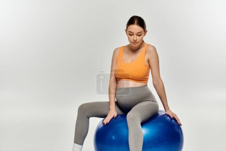 A pregnant, sporty woman balances gracefully on a large blue ball, showcasing strength and balance in an ethereal setting.