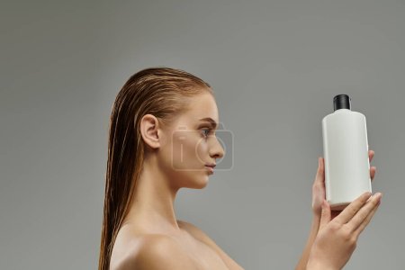 Téléchargez les photos : A young woman with wet hair delicately holds a bottle of shampoo in her hand. - en image libre de droit