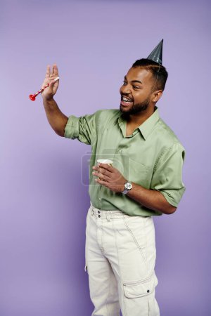 Junger afroamerikanischer Mann mit Hosenträgern hält fröhlich eine Party-Hupe in der Hand, während er einen Geburtstagshut vor einer leuchtend violetten Kulisse trägt.