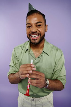 Young African American man in braces joyfully holds toothbrush while wearing party hat on purple background.