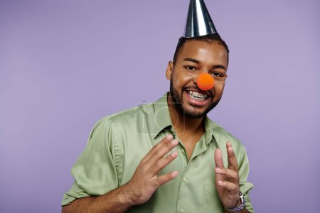 Young African American man with braces, smiling, wearing a clown nose and party hat on purple backdrop.