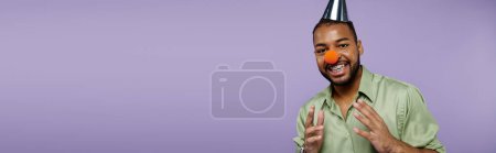 Téléchargez les photos : Un jeune Afro-Américain avec des bretelles sur fond violet équilibre joyeusement un nez de clown sur le dessus de sa tête. - en image libre de droit