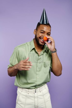 Junger Mann mit Hosenträgern setzt sich einen Partyhut auf und hält ein leuchtendes Orange vor violettem Hintergrund.