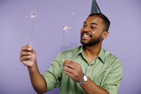 A young African American man in braces joyfully holding sparklers while wearing a party hat on a purple background.