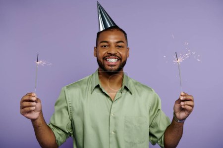 Junger schwarzer Mann in Hosenträgern lächelt und hält zwei Wunderkerzen mit Partyhut auf lila Hintergrund.