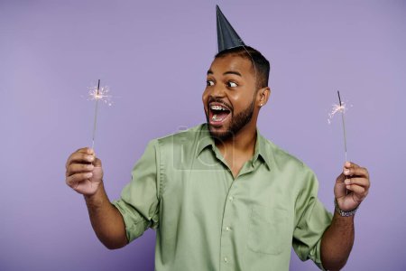 Young African American man with braces, smiling, holds two sparklers in party hat against vibrant purple background.