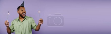 Junger afroamerikanischer Mann mit Zahnspange balanciert auf zwei Wunderkerzen und trägt einen festlichen Partyhut auf lila Hintergrund.