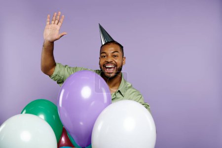 Joven hombre afroamericano en sombrero de fiesta sonriendo, sosteniendo globos de colores sobre un fondo púrpura.