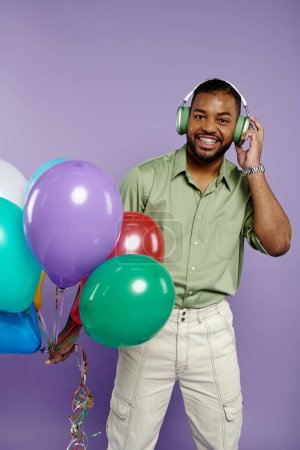 Jeune Afro-Américain aux accolades souriantes, tenant ballons et écouteurs sur fond violet.