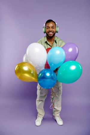 Un homme afro-américain élégant aux bretelles tient un bouquet coloré de ballons et d'écouteurs à la mode sur fond violet.