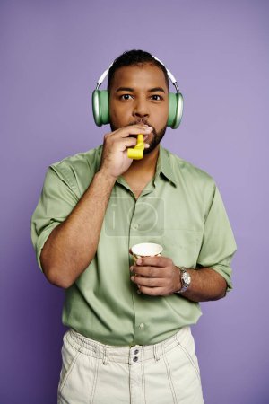 Young African American man joyfully listens to music while blowing party horn against a purple backdrop.