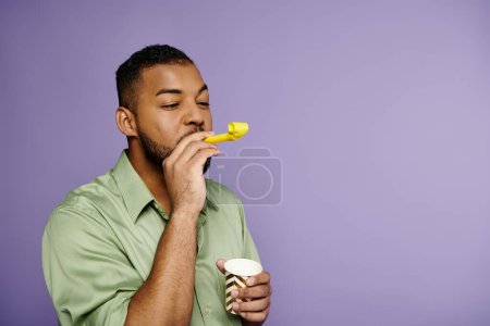 A young African American man in a green shirt blowing party horn on a purple background.