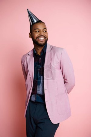 Young, happy African American man in pink jacket and party hat on pink background, full of joy and excitement.