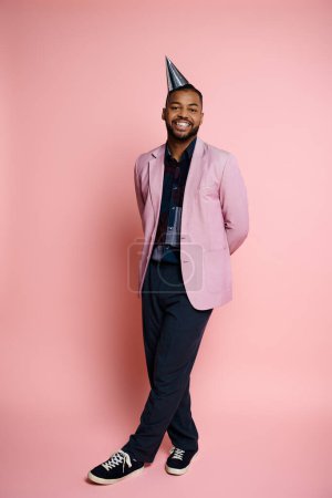 A stylish young African American man in a pink suit and party hat, exuding happiness on a vibrant pink background.