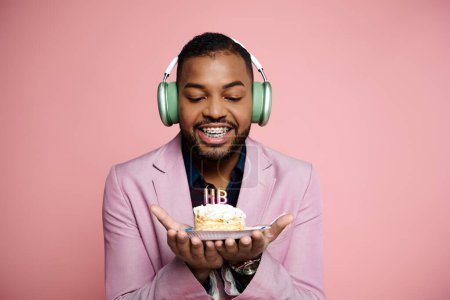 Téléchargez les photos : Joyeux jeune Afro-Américain en bretelles écoute de la musique sur casque tout en tenant un gâteau sur fond rose. - en image libre de droit
