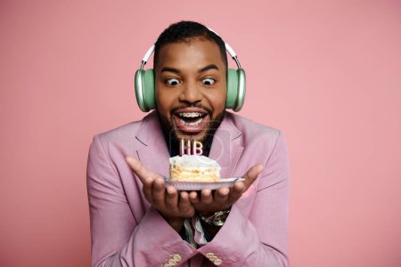 Young African American man with braces happily holds a piece of cake while wearing headphones on a pink background.