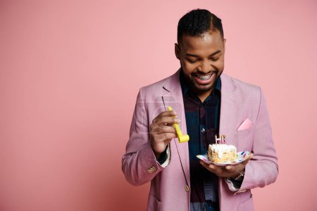 Ein stilvoller junger Afroamerikaner im rosafarbenen Anzug hält glücklich einen Teller mit Geburtstagstorte und Partyhorn vor einem leuchtend rosafarbenen Hintergrund.