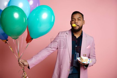 Ein junger afroamerikanischer Mann in Hosenträgern hält einen Teller mit Kuchen und Luftballons in der Hand und lächelt zur Feier auf rosa Hintergrund.