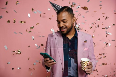 Young African American man in braces, happy, holding cup and cell phone, standing in party hat on a pink background.