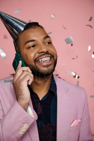 Téléchargez les photos : Jeune homme afro-américain heureux avec des bretelles, portant un chapeau de fête, parlant sur un téléphone portable sur un fond rose. - en image libre de droit