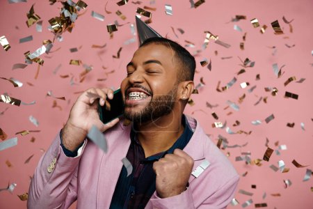 Junger afroamerikanischer Mann in Hosenträgern trägt einen Partyhut und spricht fröhlich auf einem Handy vor rosa Hintergrund.