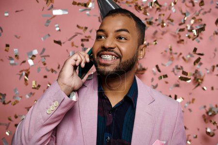 Young, joyful African American man wearing a party hat conversing on a cell phone on a pink background.
