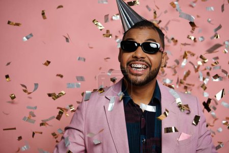 Photo for Young African American man joyfully wearing a party hat and sunglasses, exuding happiness on a pink background. - Royalty Free Image