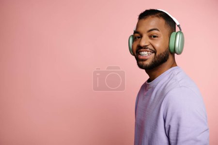 Téléchargez les photos : Jeune homme afro-américain avec des bretelles souriant vivement tout en portant des écouteurs sur un fond rose. - en image libre de droit