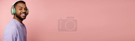 Téléchargez les photos : Vibrant jeune homme afro-américain en chemise violette et écouteurs sourit sur fond rose. - en image libre de droit