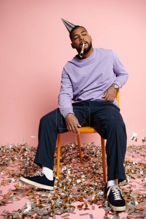 Young African American man with braces wearing a party hat sitting on a chair against a pink background.