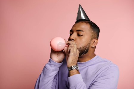 Young African American man happily blows a large bubble while wearing a party hat on a vibrant pink background.